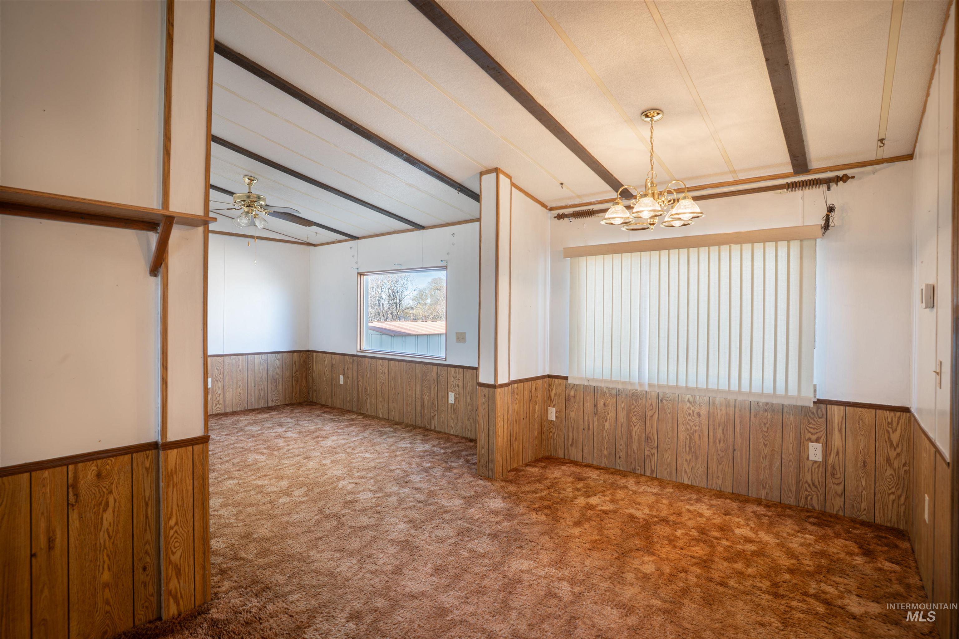 Carpeted empty room with wood walls, a wainscoted wall, beamed ceiling, a ceiling fan, and a chandelier