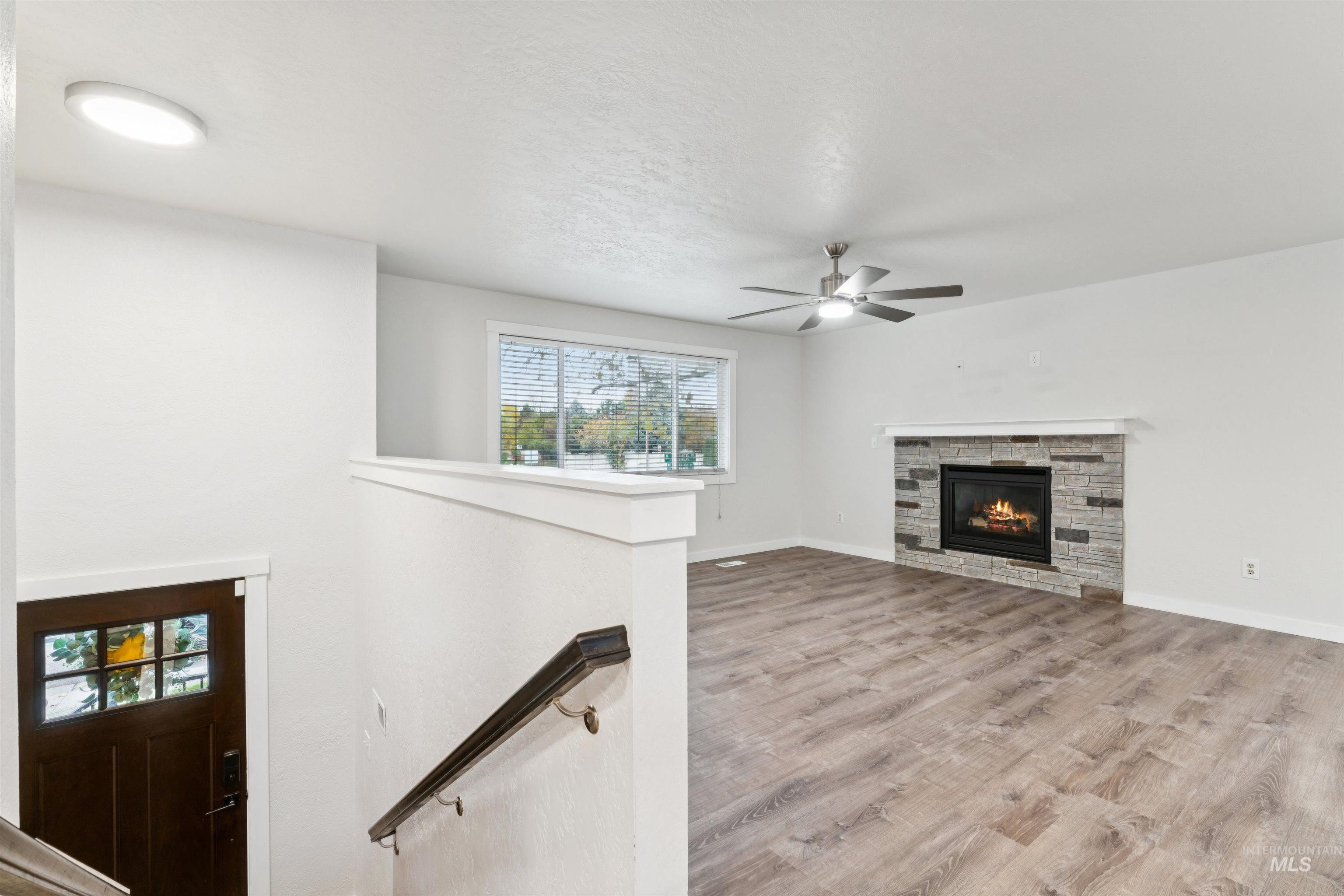Unfurnished living room with wood finished floors, a stone fireplace, a ceiling fan, and a textured ceiling