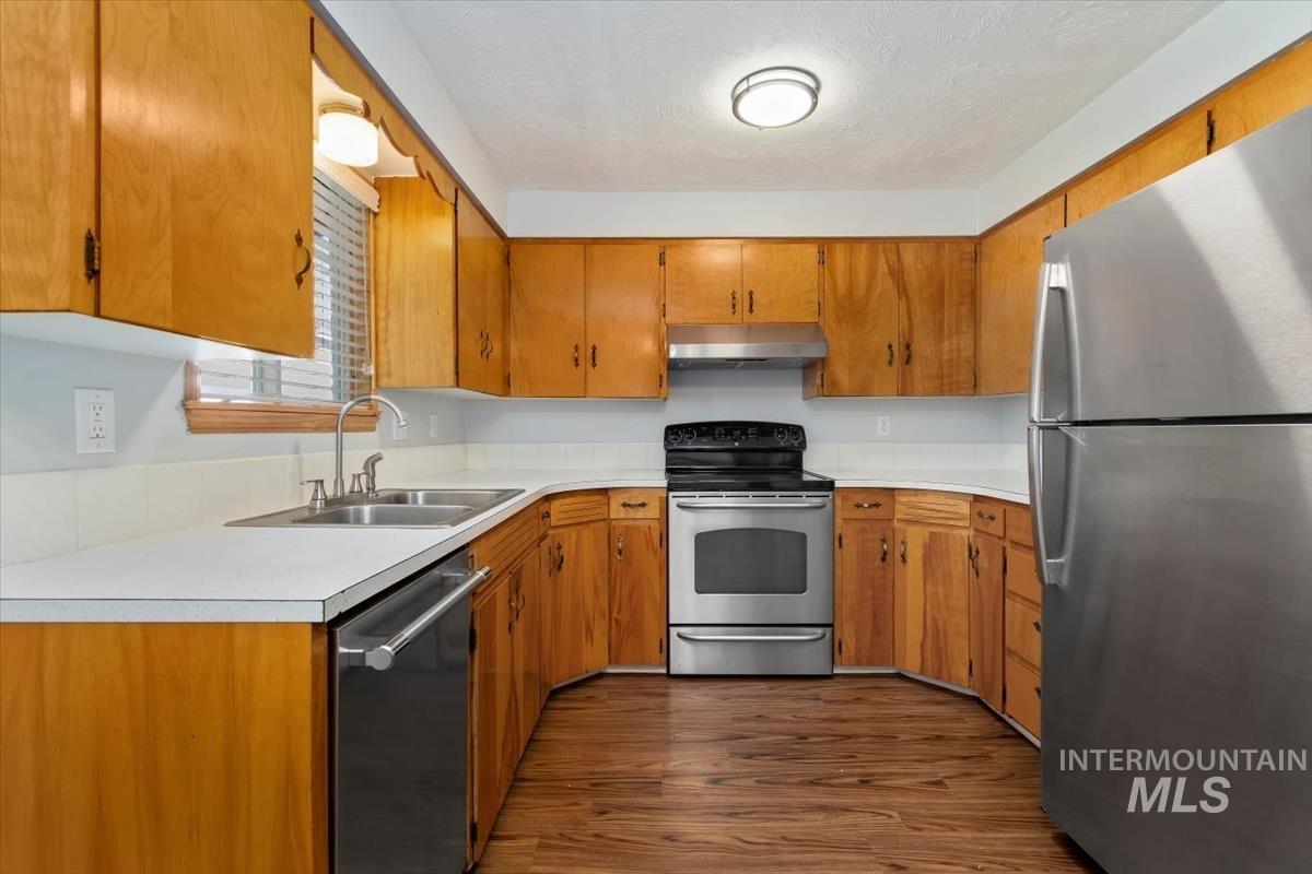 Kitchen with appliances with stainless steel finishes, light countertops, brown cabinetry, dark wood finished floors, and under cabinet range hood