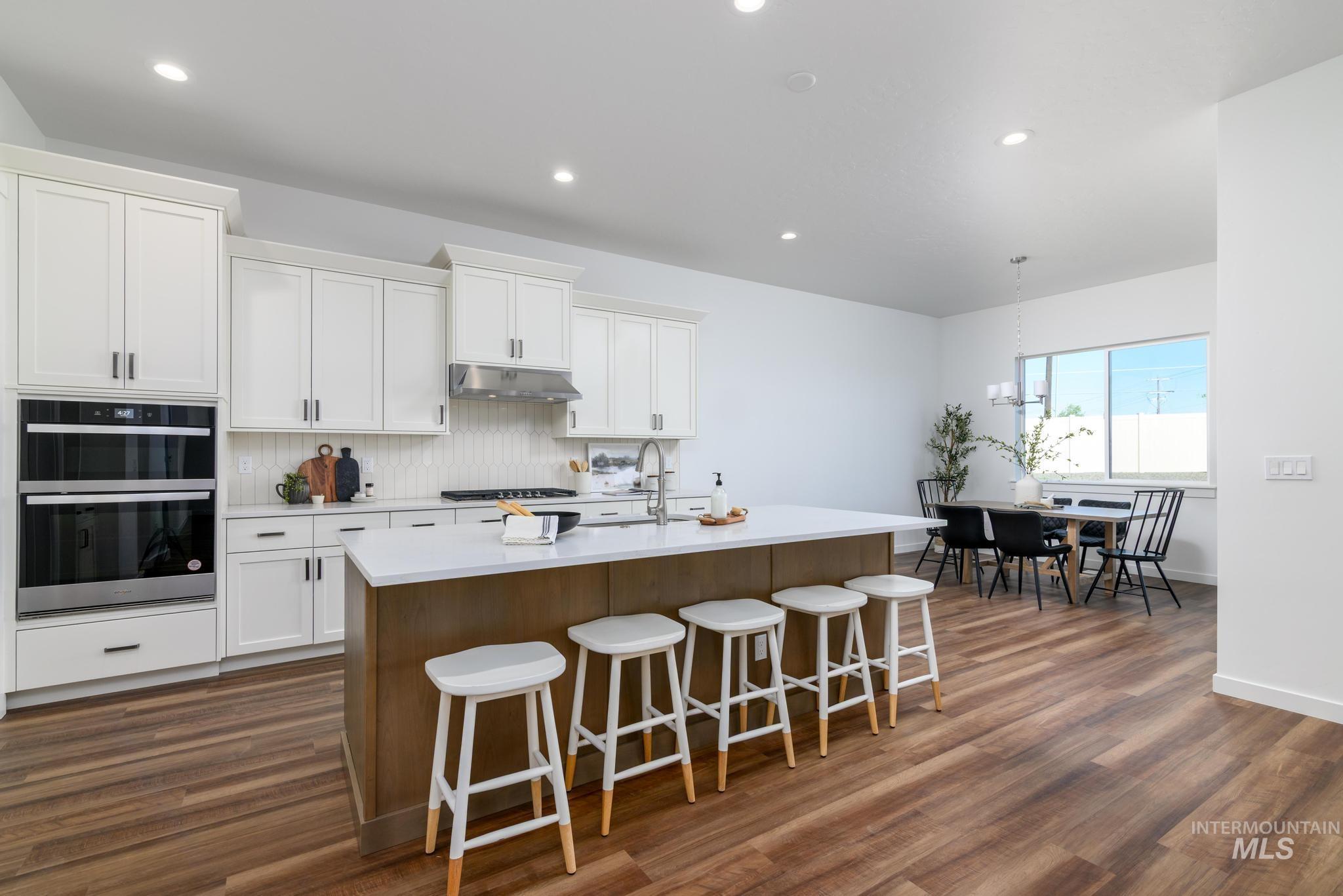 Kitchen with under cabinet range hood, double oven, light countertops, an island with sink, and decorative backsplash