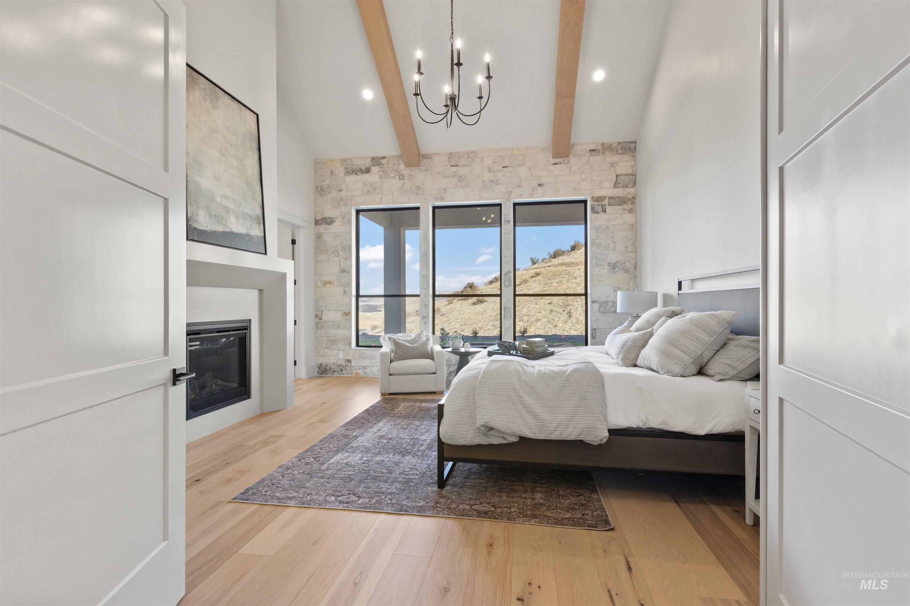 Bedroom with light wood-style floors, a glass covered fireplace, a chandelier, beamed ceiling, and a high ceiling