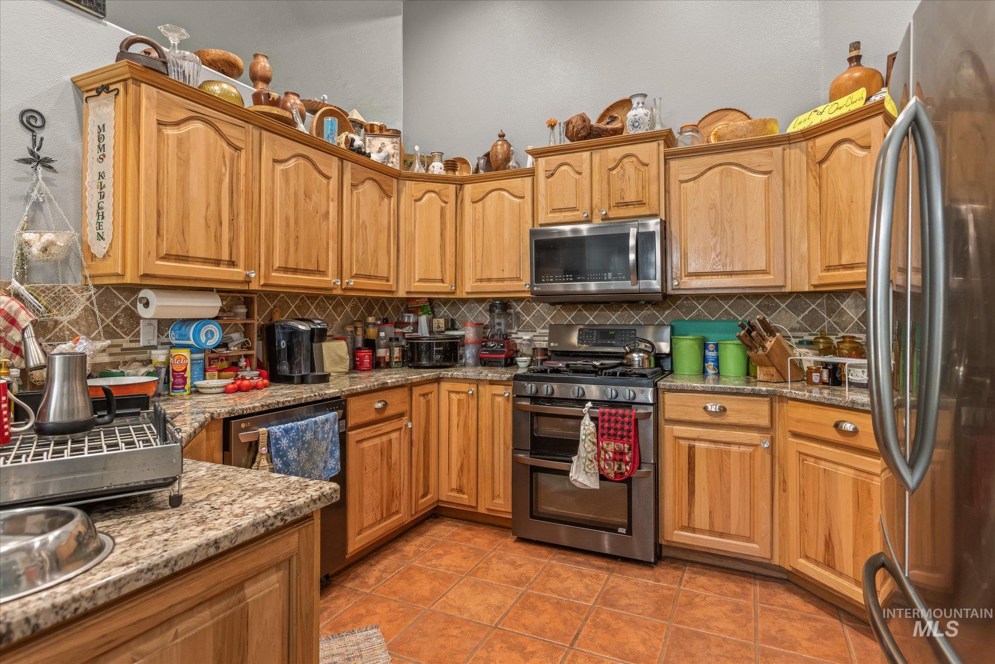 Kitchen featuring stainless steel appliances, light stone countertops, decorative backsplash, light tile patterned floors, and brown cabinets
