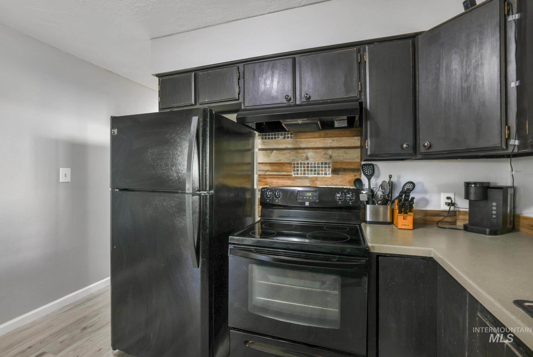 Kitchen featuring black appliances, dark cabinets, light wood-style floors, ventilation hood, and light countertops