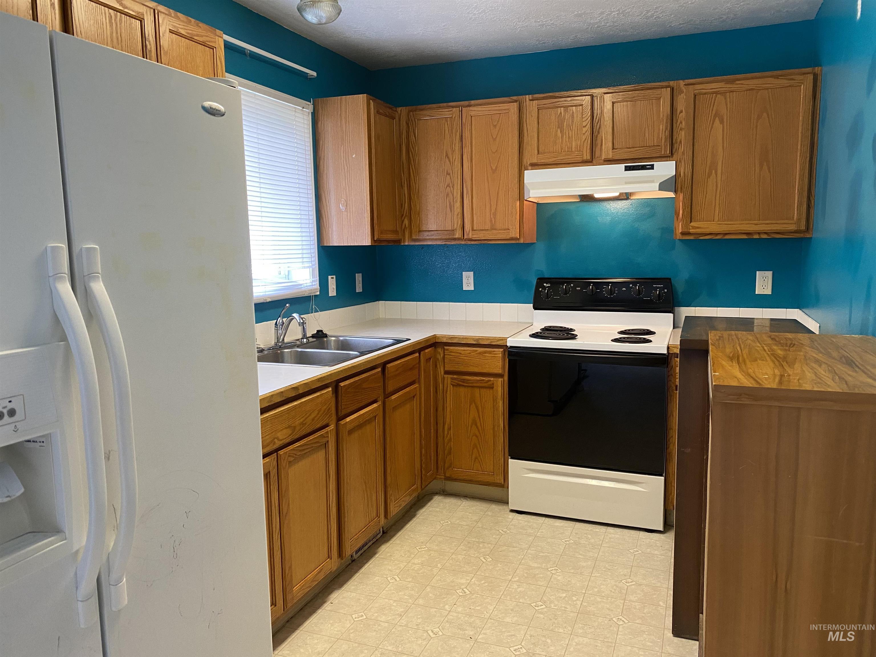 Kitchen featuring white fridge with ice dispenser, range with electric stovetop, brown cabinets, under cabinet range hood, and light floors