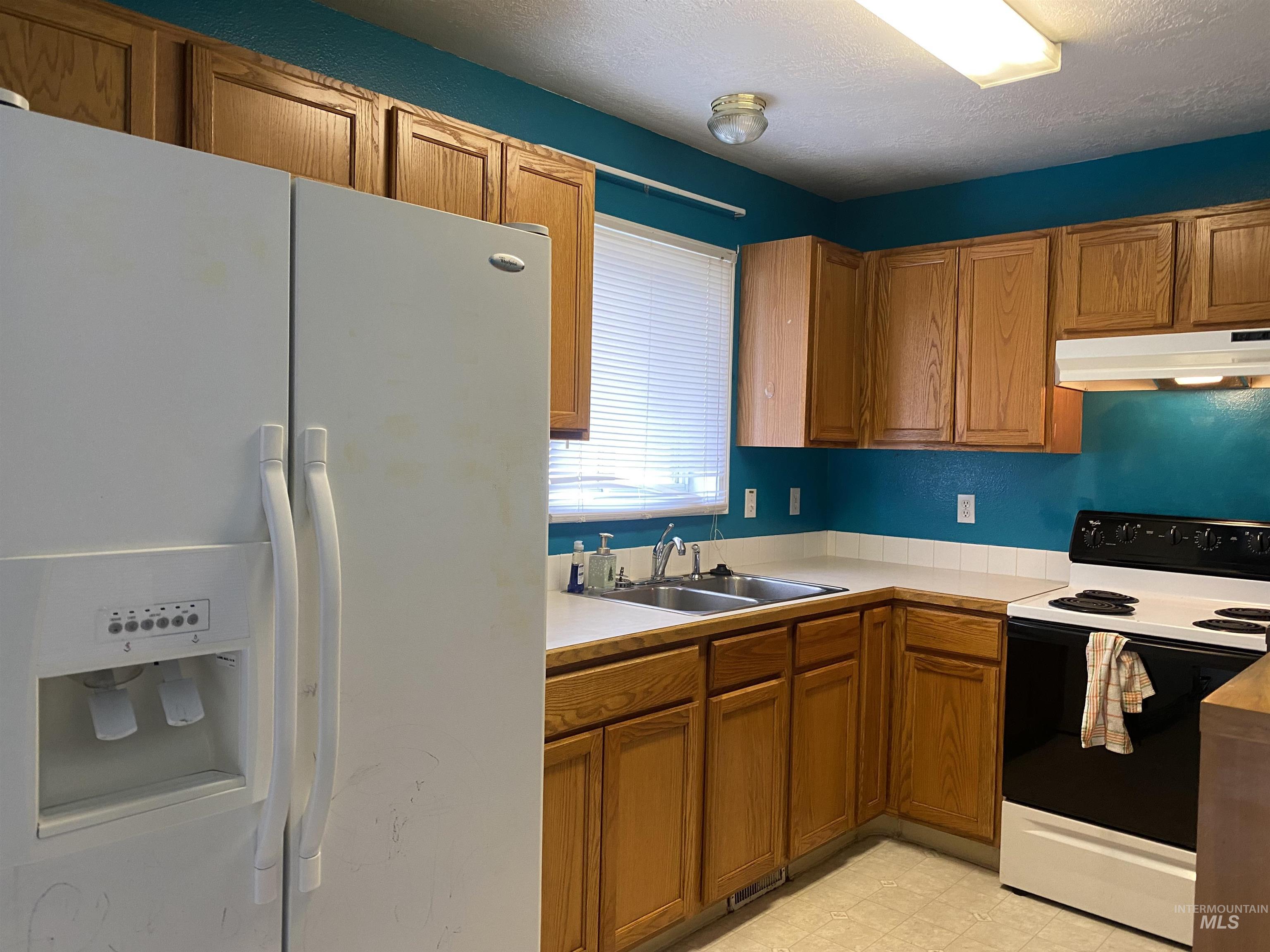 Kitchen with white appliances, brown cabinetry, light countertops, under cabinet range hood, and a textured ceiling
