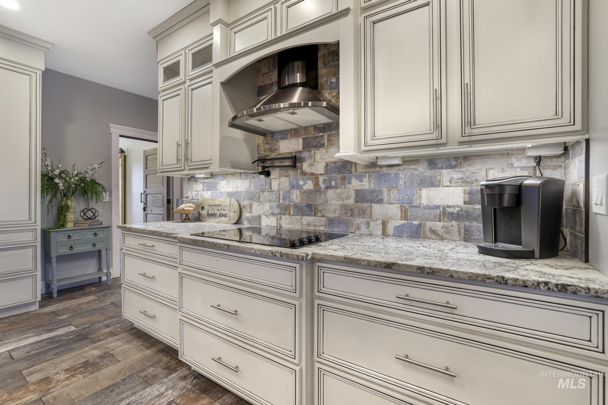 Kitchen with wall chimney range hood, decorative backsplash, light stone counters, glass insert cabinets, and dark wood finished floors