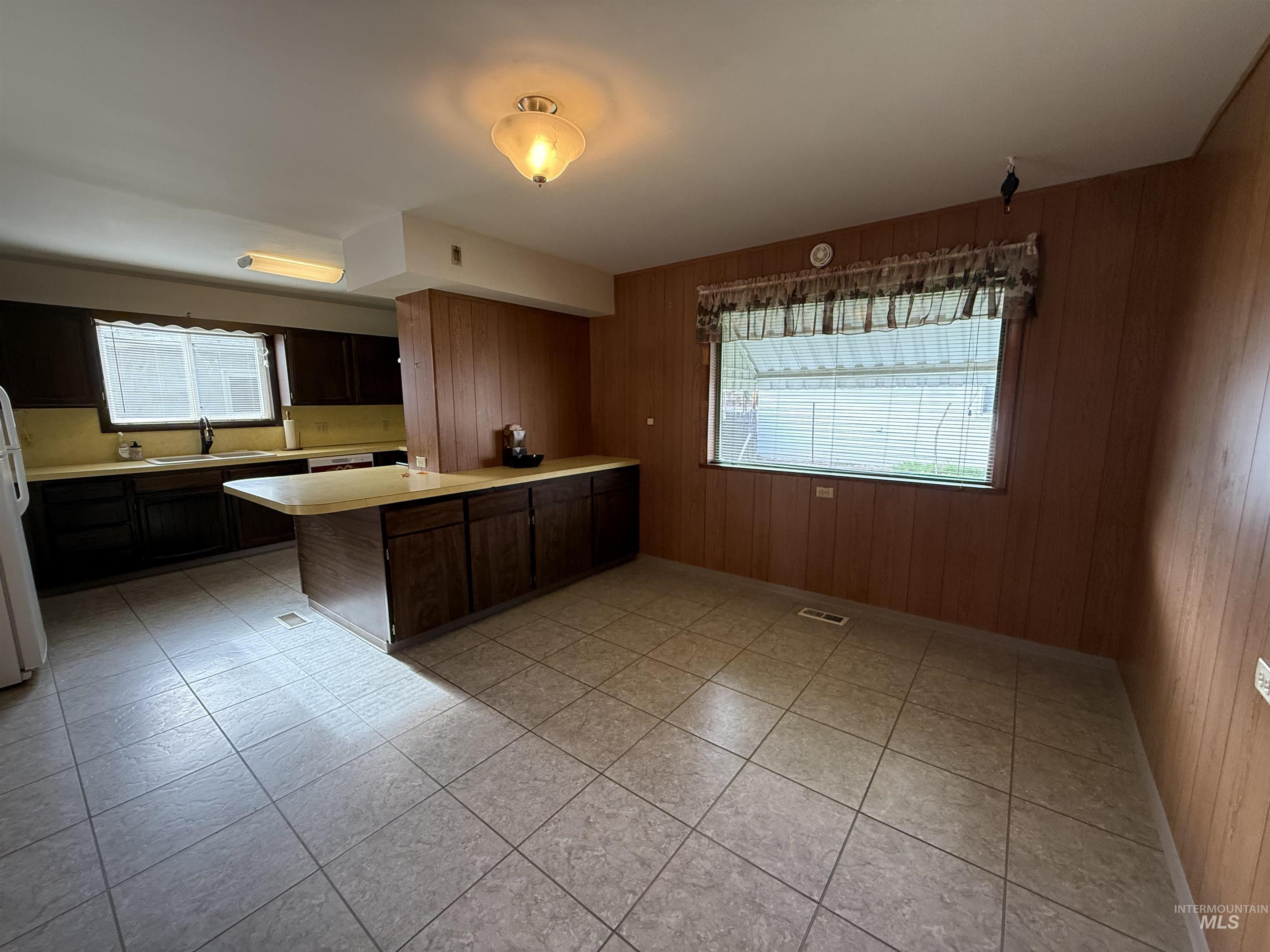 Kitchen with light countertops, a peninsula, wood walls, dark brown cabinets, and freestanding refrigerator