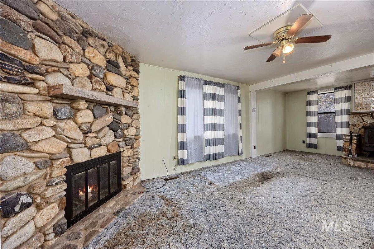 Unfurnished living room featuring a stone fireplace, carpet floors, a textured ceiling, and a ceiling fan