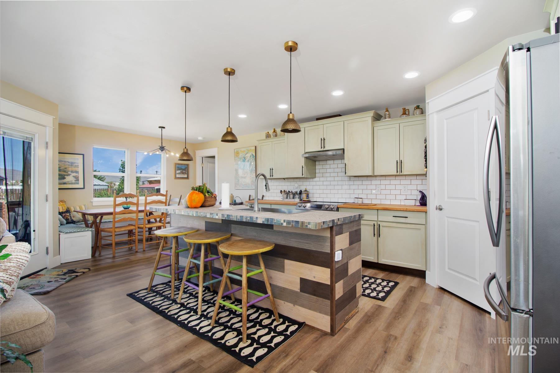 Kitchen featuring freestanding refrigerator, tasteful backsplash, decorative light fixtures, an island with sink, and light wood-type flooring