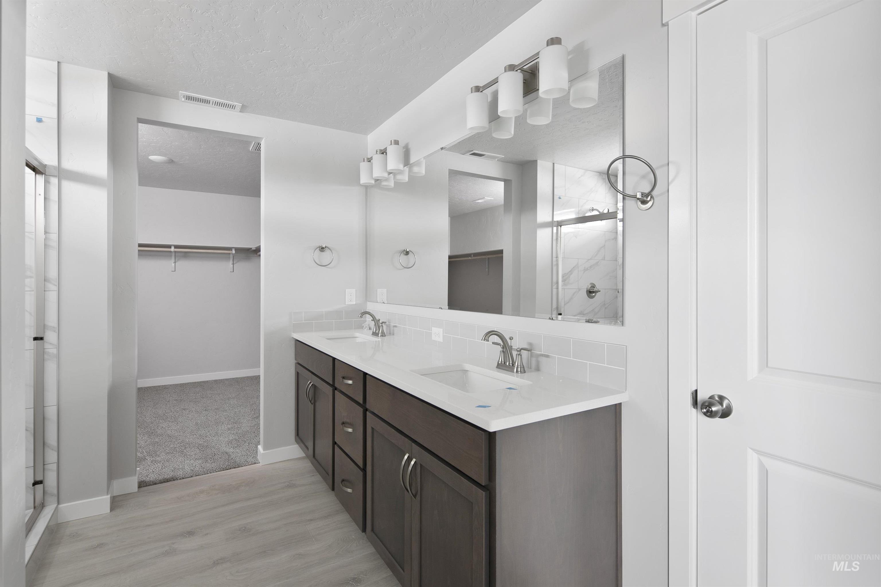 Full bath with a spacious closet, a shower stall, double vanity, a textured ceiling, and light wood finished floors