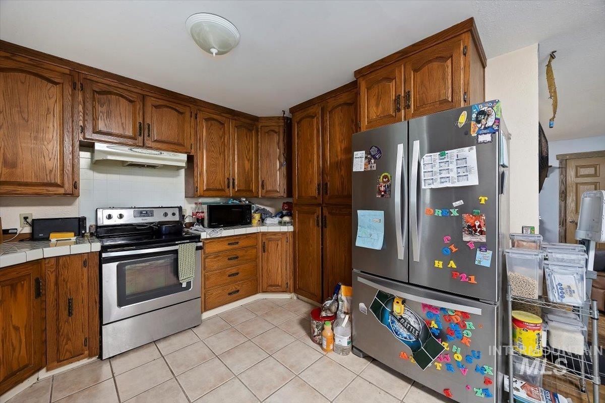 Kitchen featuring tile countertops, stainless steel appliances, decorative backsplash, light tile patterned flooring, and brown cabinets
