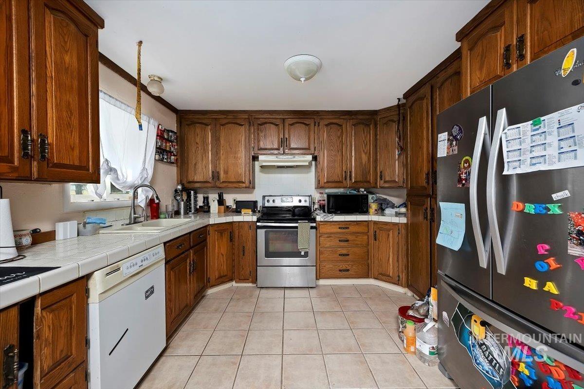 Kitchen featuring stainless steel appliances, light tile patterned floors, tile countertops, brown cabinets, and under cabinet range hood