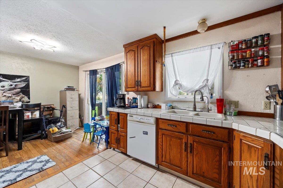 Kitchen featuring a textured wall, tile counters, light tile patterned flooring, brown cabinetry, and white dishwasher
