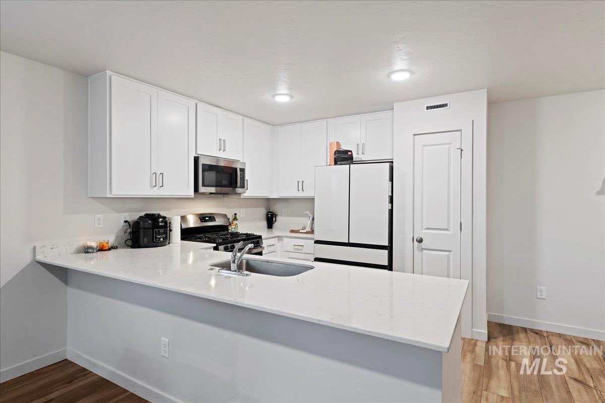 Kitchen with white cabinetry, appliances with stainless steel finishes, a peninsula, light stone countertops, and light wood-type flooring