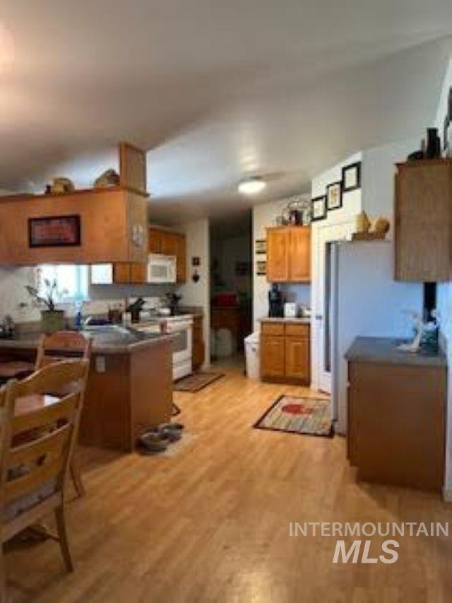 Kitchen with brown cabinets, light wood finished floors, white appliances, dark countertops, and a peninsula