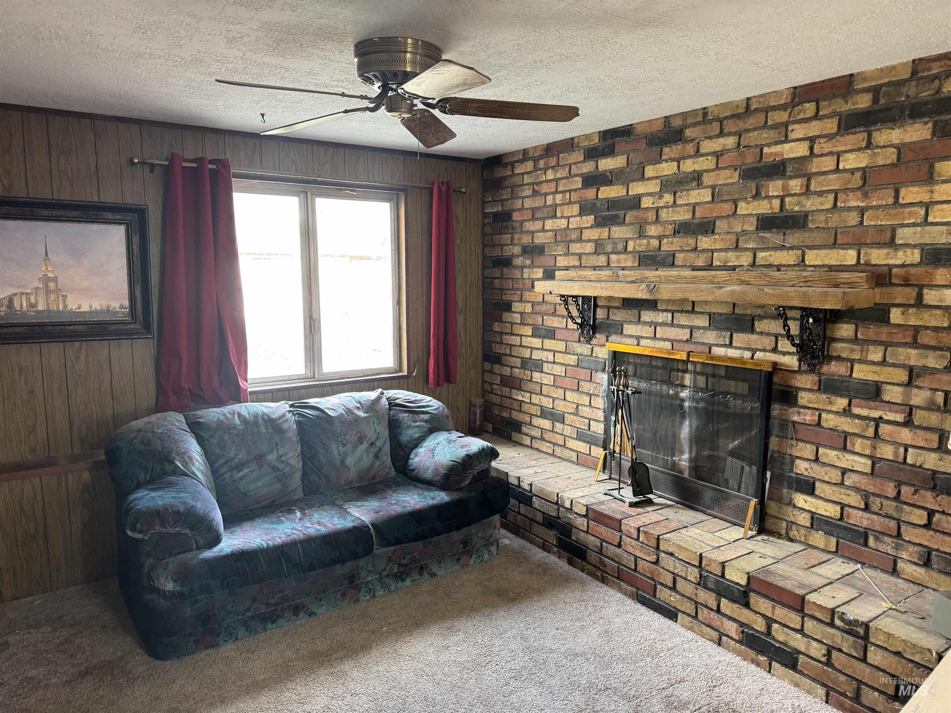 Carpeted living room with a textured ceiling, a brick fireplace, wood walls, and a ceiling fan