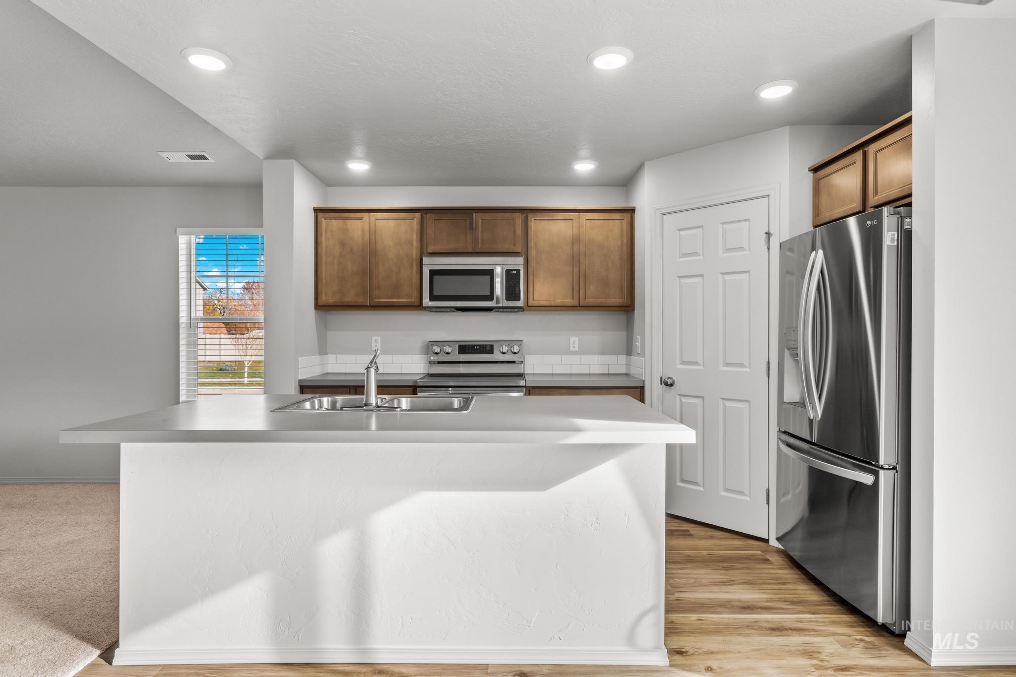 Kitchen with stainless steel appliances, recessed lighting, light countertops, a center island with sink, and brown cabinetry