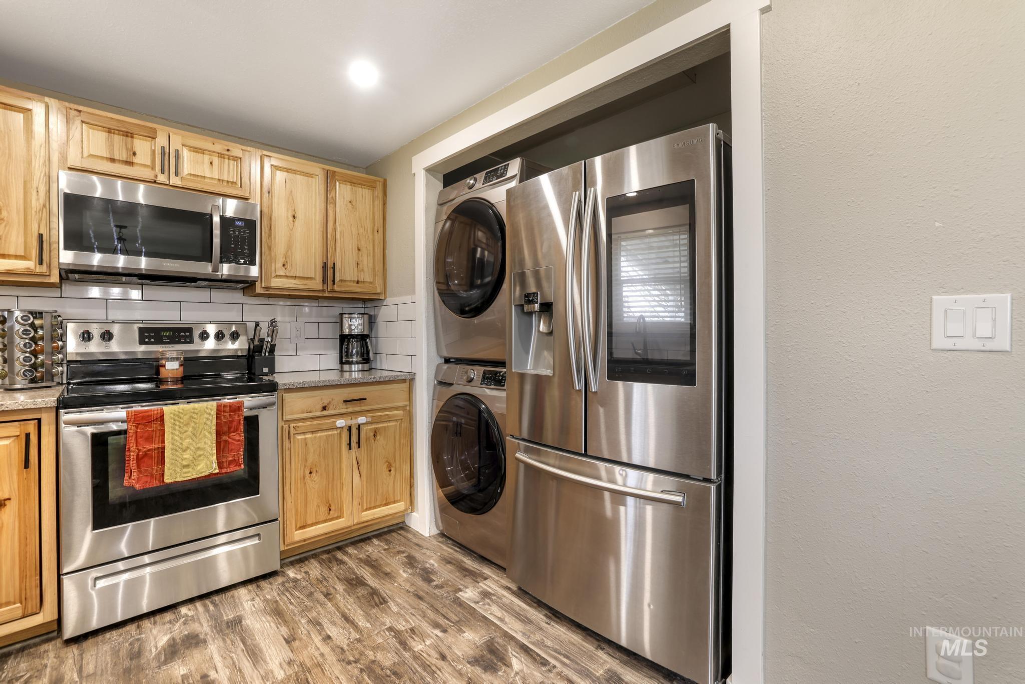 Kitchen with appliances with stainless steel finishes, decorative backsplash, dark wood-type flooring, light stone counters, and stacked washer and clothes dryer