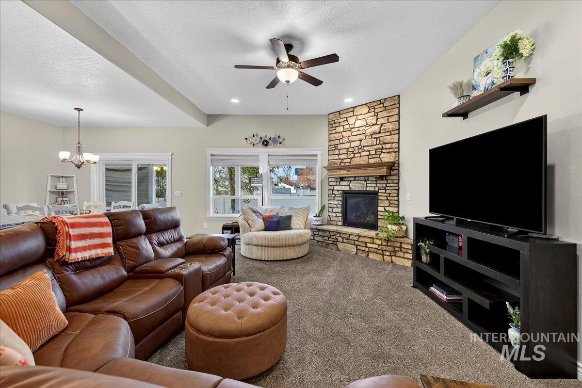 Carpeted living room with a textured ceiling, a stone fireplace, a ceiling fan, a chandelier, and recessed lighting