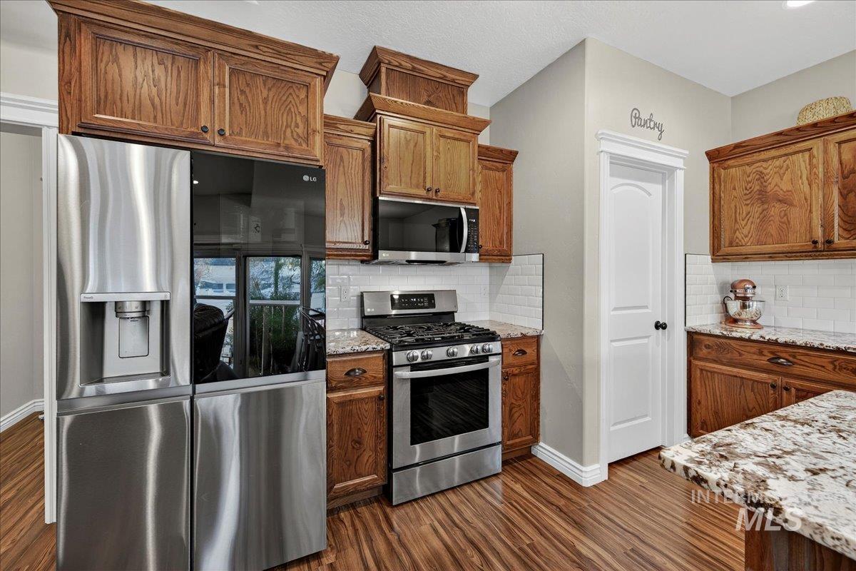 Kitchen with appliances with stainless steel finishes, dark wood-style flooring, brown cabinetry, light stone counters, and backsplash