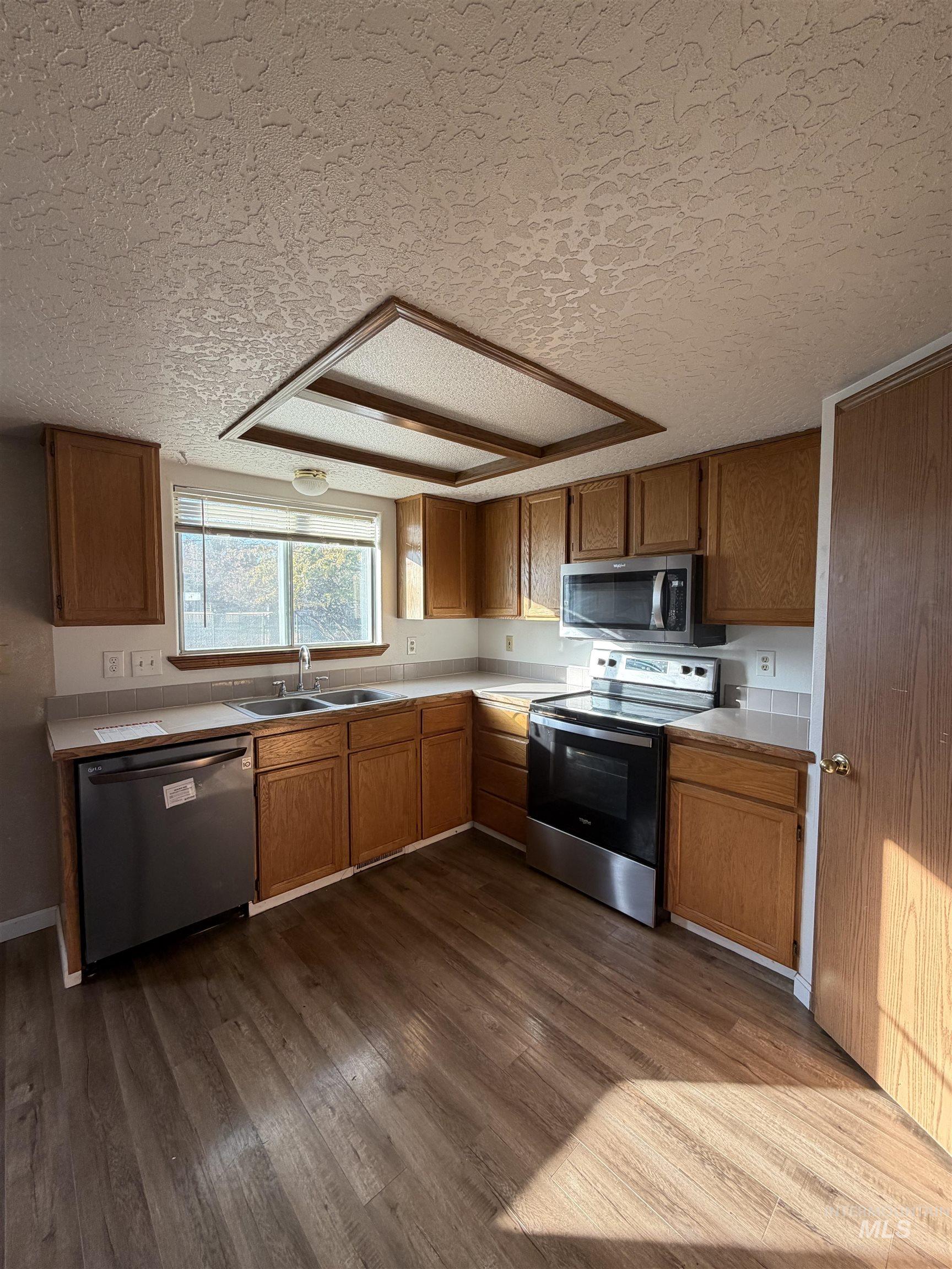 Kitchen featuring light countertops, appliances with stainless steel finishes, a textured ceiling, dark wood-style floors, and brown cabinetry