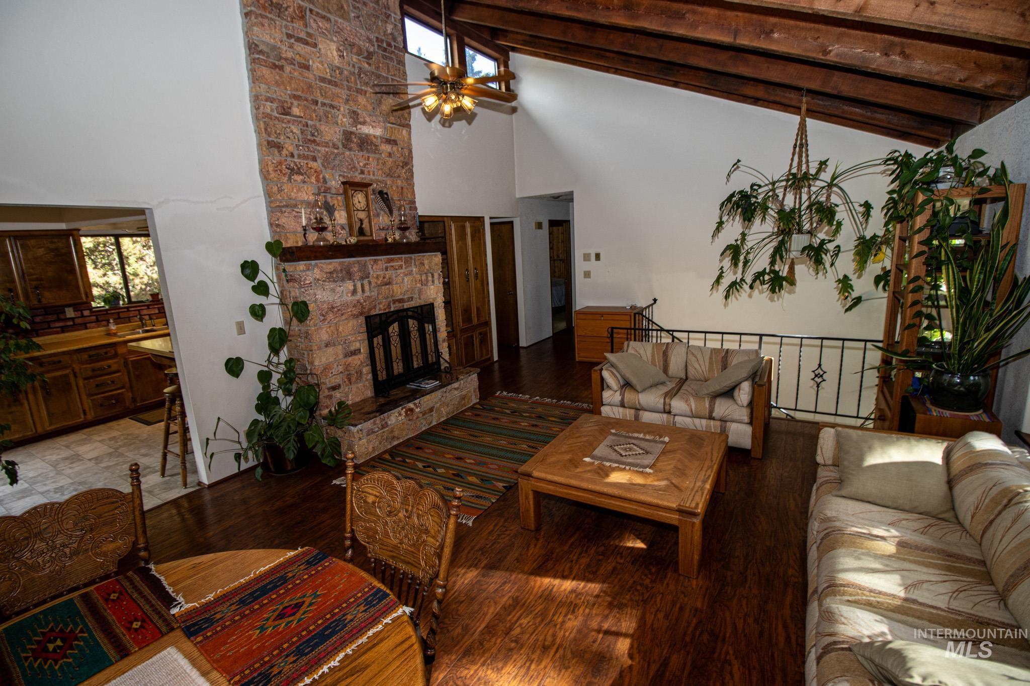 Living room with beam ceiling, high vaulted ceiling, ceiling fan, a stone fireplace, and wood finished floors