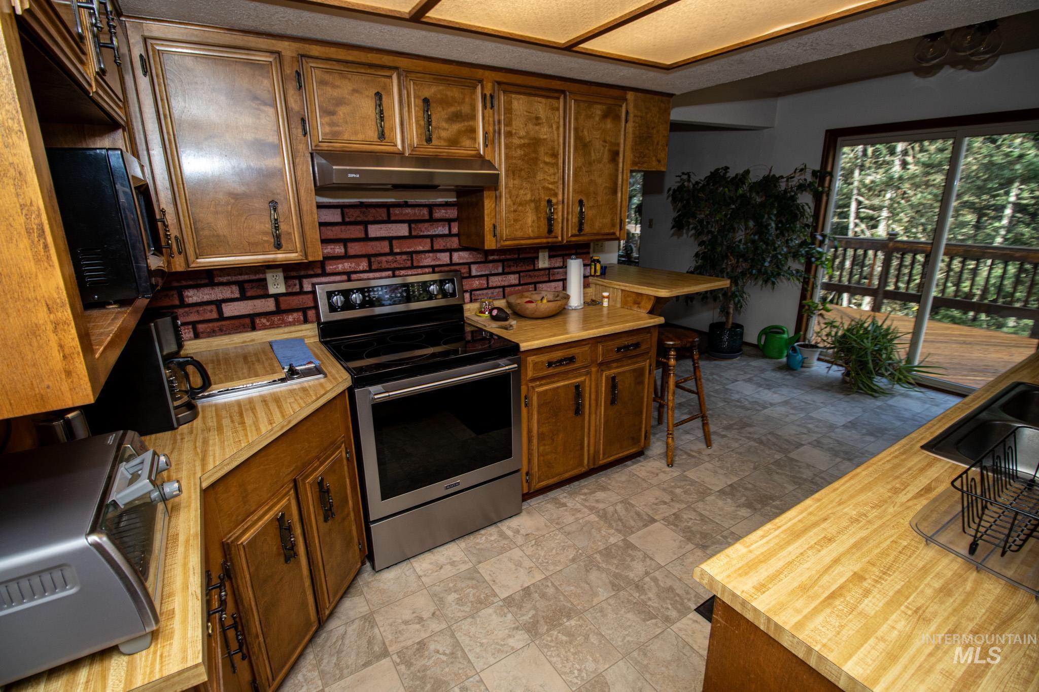 Kitchen with stainless steel range with electric cooktop, brown cabinetry, under cabinet range hood, black microwave, and decorative backsplash