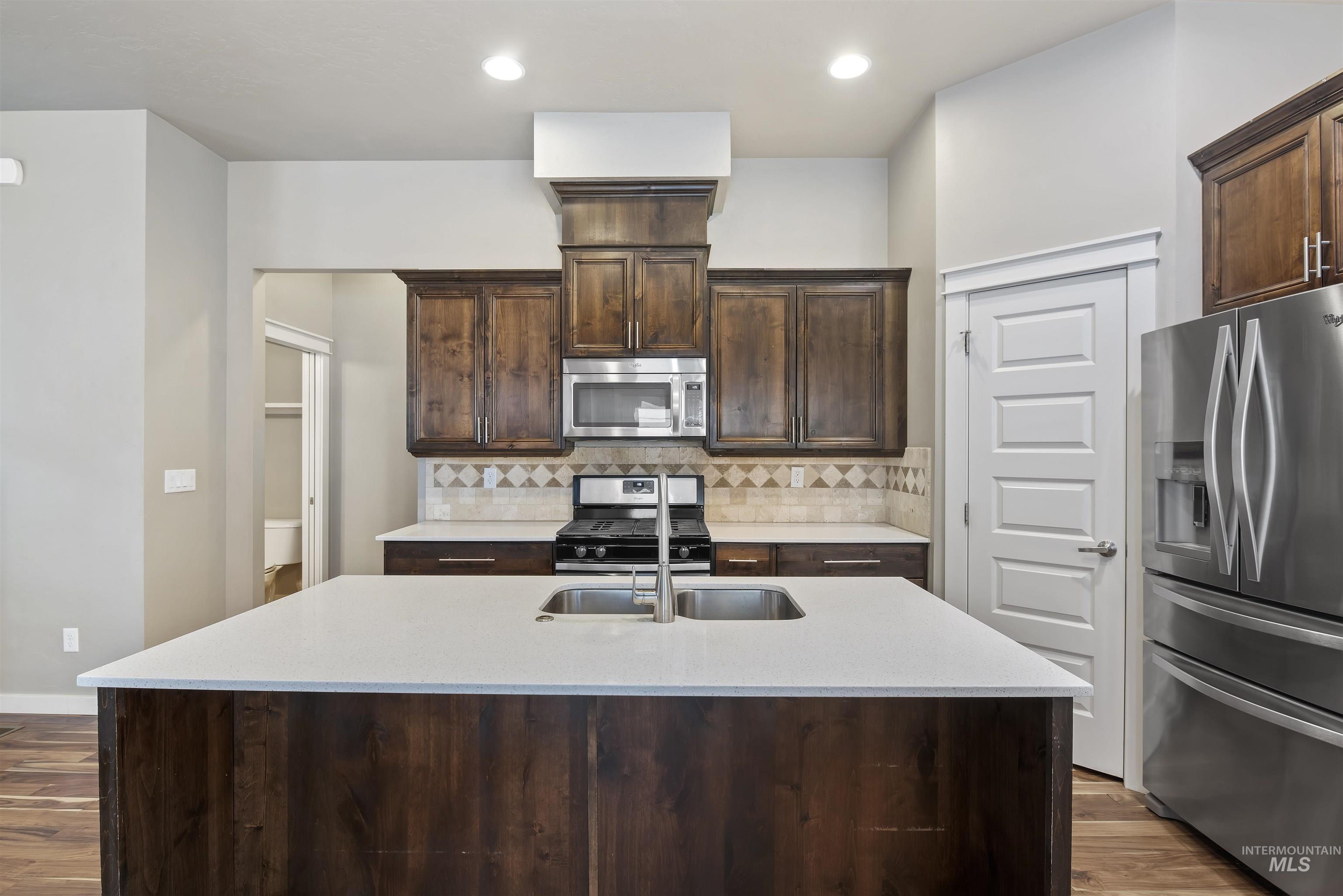 Kitchen with appliances with stainless steel finishes, tasteful backsplash, light stone countertops, a kitchen island with sink, and dark wood finished floors