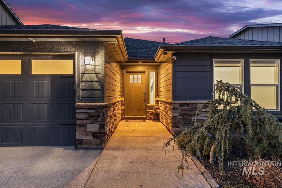View of exterior entry with stone siding, an attached garage, roof with shingles, and driveway