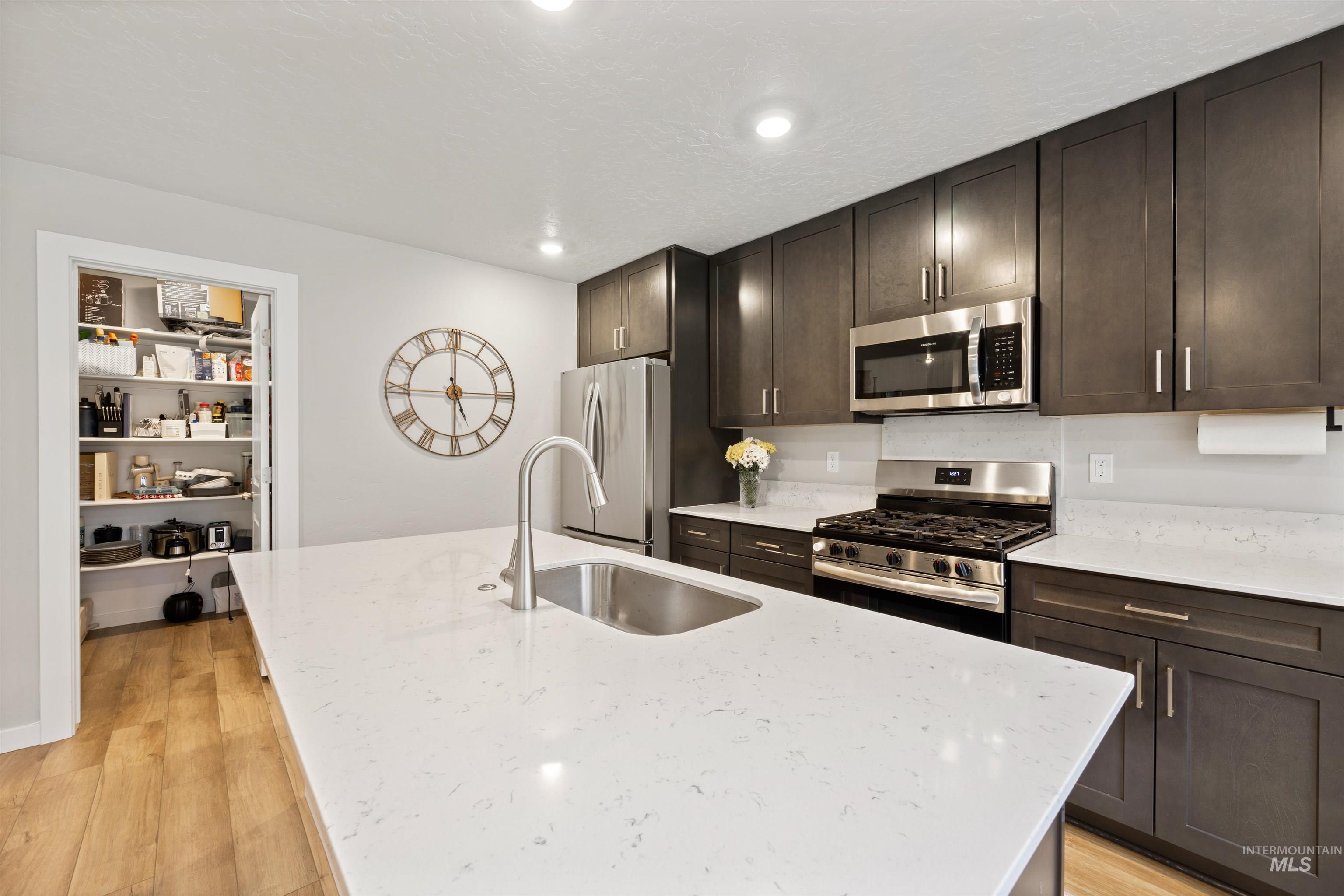 Kitchen with stainless steel appliances, light wood-type flooring, dark brown cabinetry, light stone countertops, and a center island with sink
