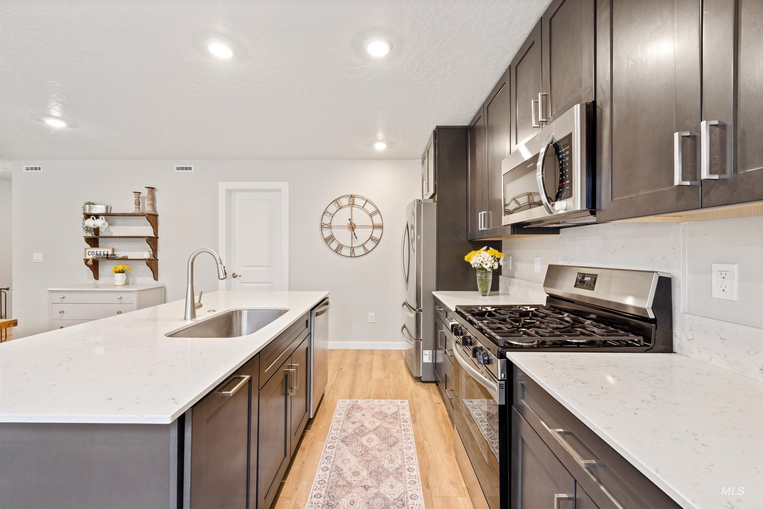 Kitchen with dark brown cabinets, appliances with stainless steel finishes, light wood finished floors, light stone countertops, and recessed lighting