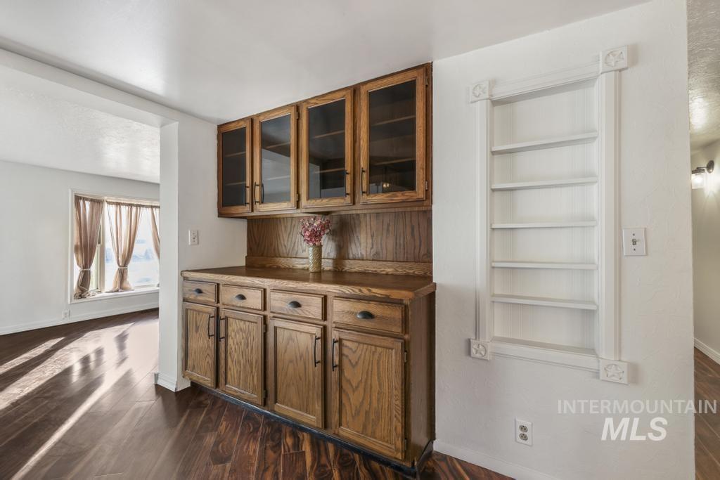 Bar featuring dark wood-style flooring, brown cabinetry, built in features, dark countertops, and glass insert cabinets