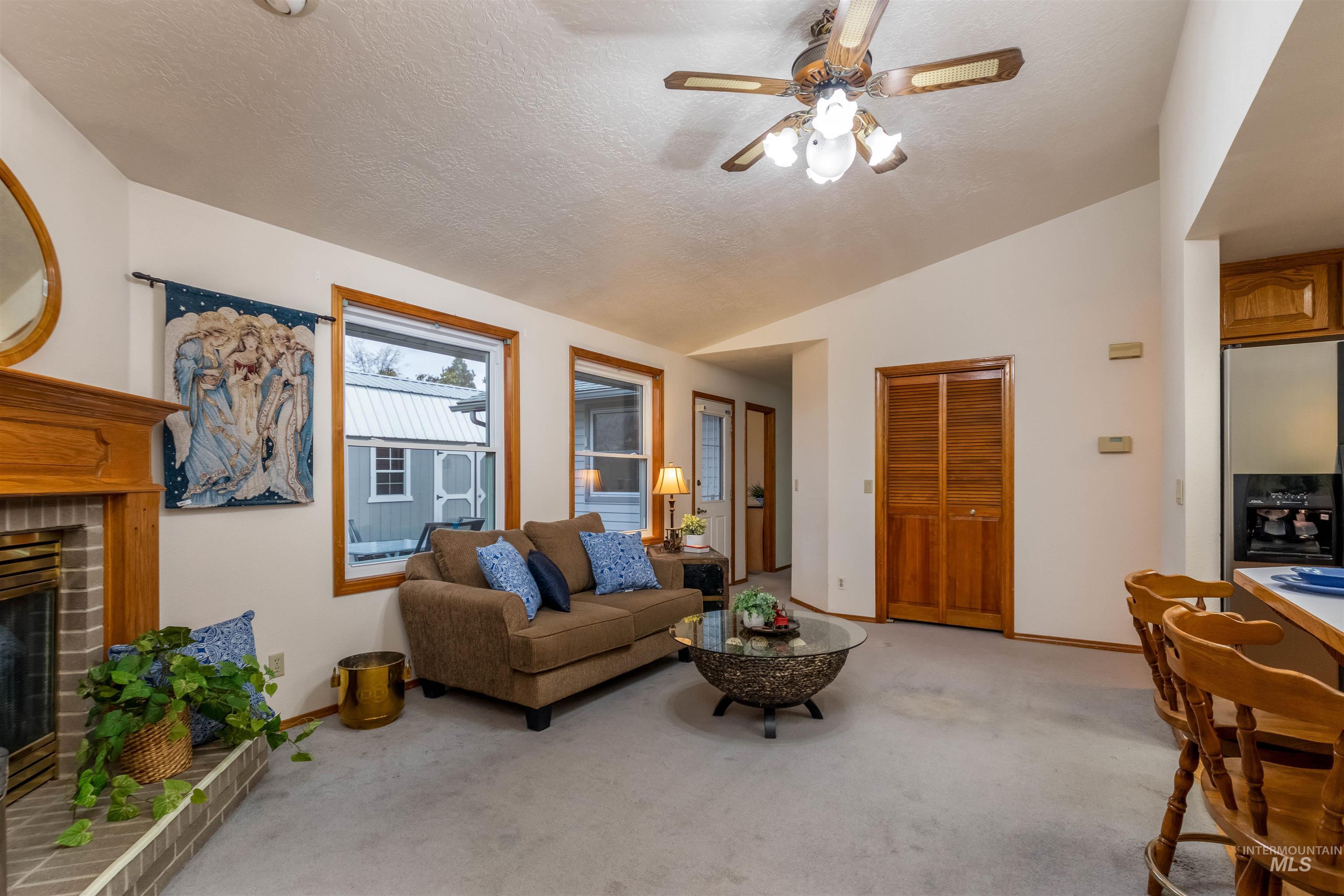 Living area featuring vaulted ceiling, a fireplace, light carpet, a textured ceiling, and a ceiling fan