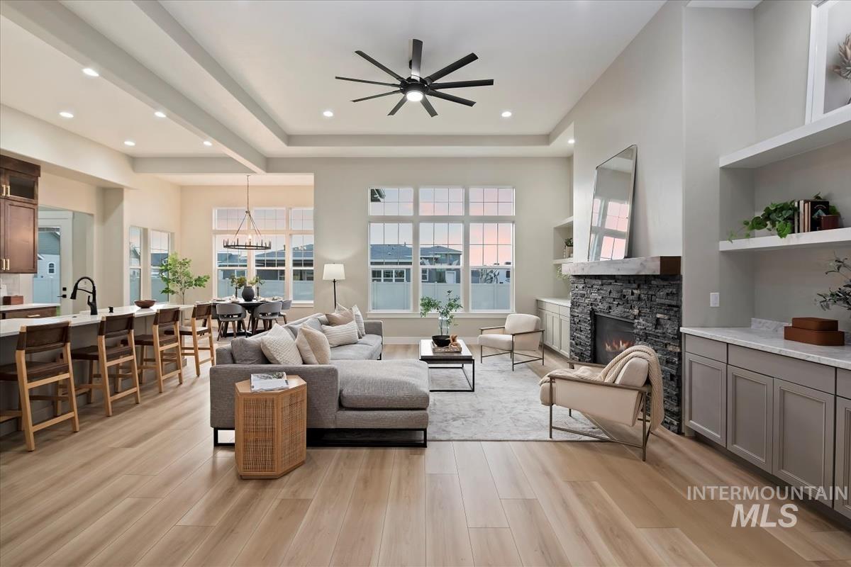 Living area with light wood-type flooring, a stone fireplace, a ceiling fan, a chandelier, and a tray ceiling
