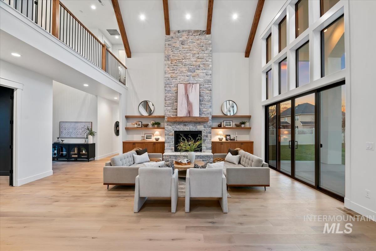 Living room featuring a towering ceiling, a stone fireplace, light wood finished floors, recessed lighting, and beam ceiling