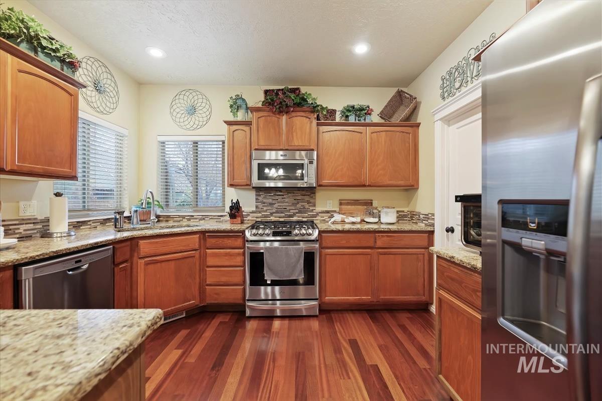 Kitchen featuring appliances with stainless steel finishes, dark wood-style flooring, light stone counters, brown cabinets, and recessed lighting