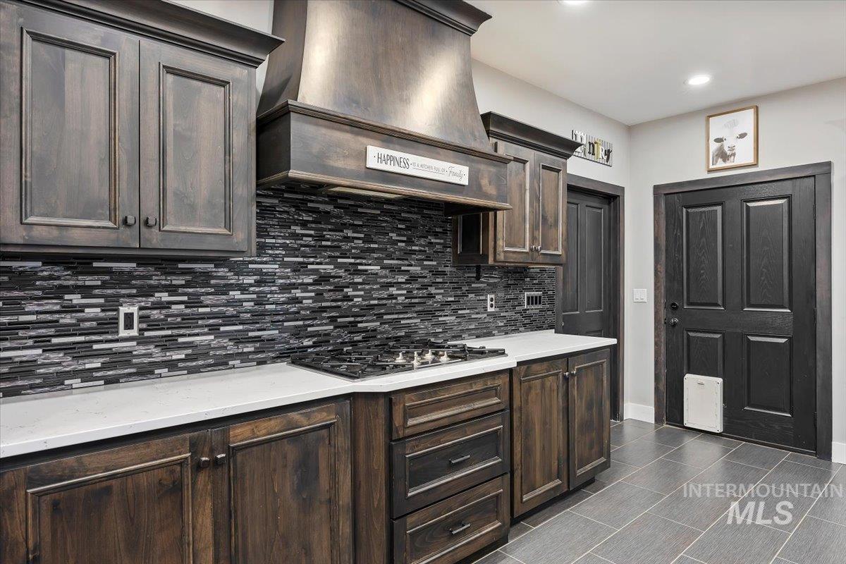 Kitchen featuring dark brown cabinetry, premium range hood, stainless steel gas cooktop, tasteful backsplash, and light stone counters