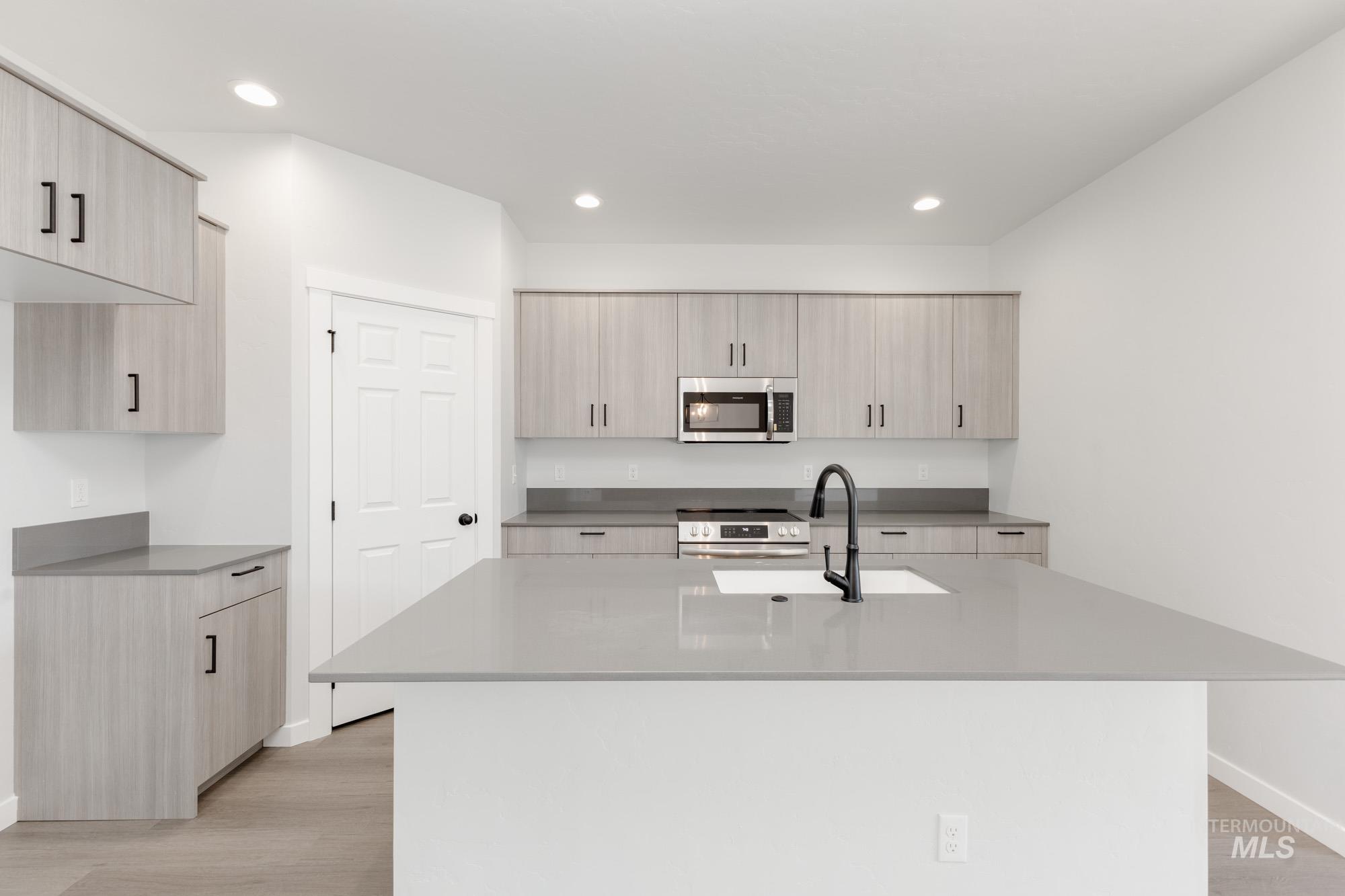 Kitchen featuring appliances with stainless steel finishes, light wood finished floors, light brown cabinets, dark stone counters, and a kitchen island with sink