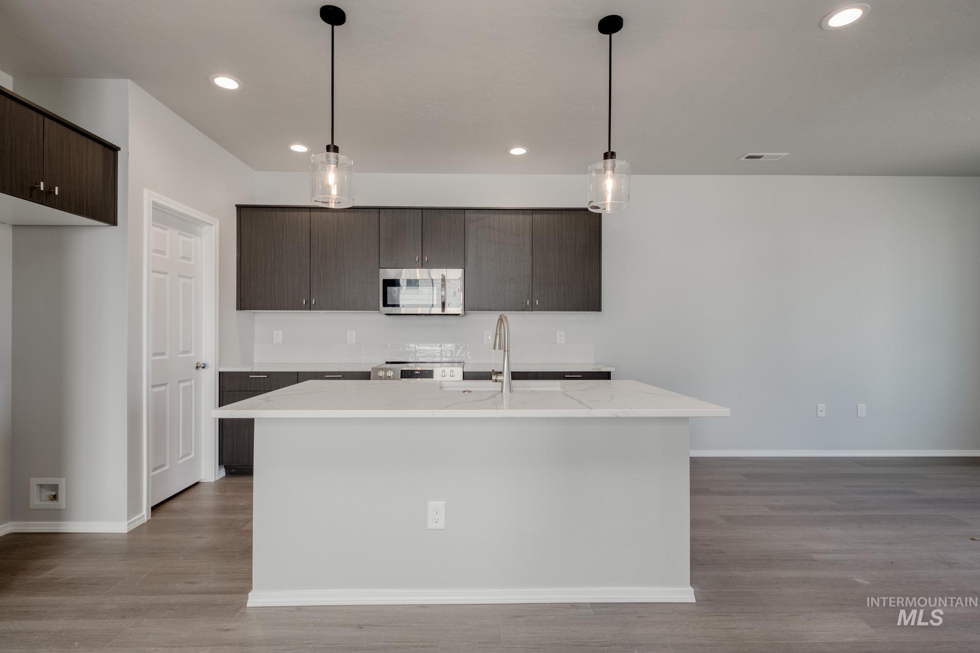 Kitchen with dark brown cabinetry, decorative light fixtures, stainless steel appliances, a kitchen island with sink, and light stone countertops