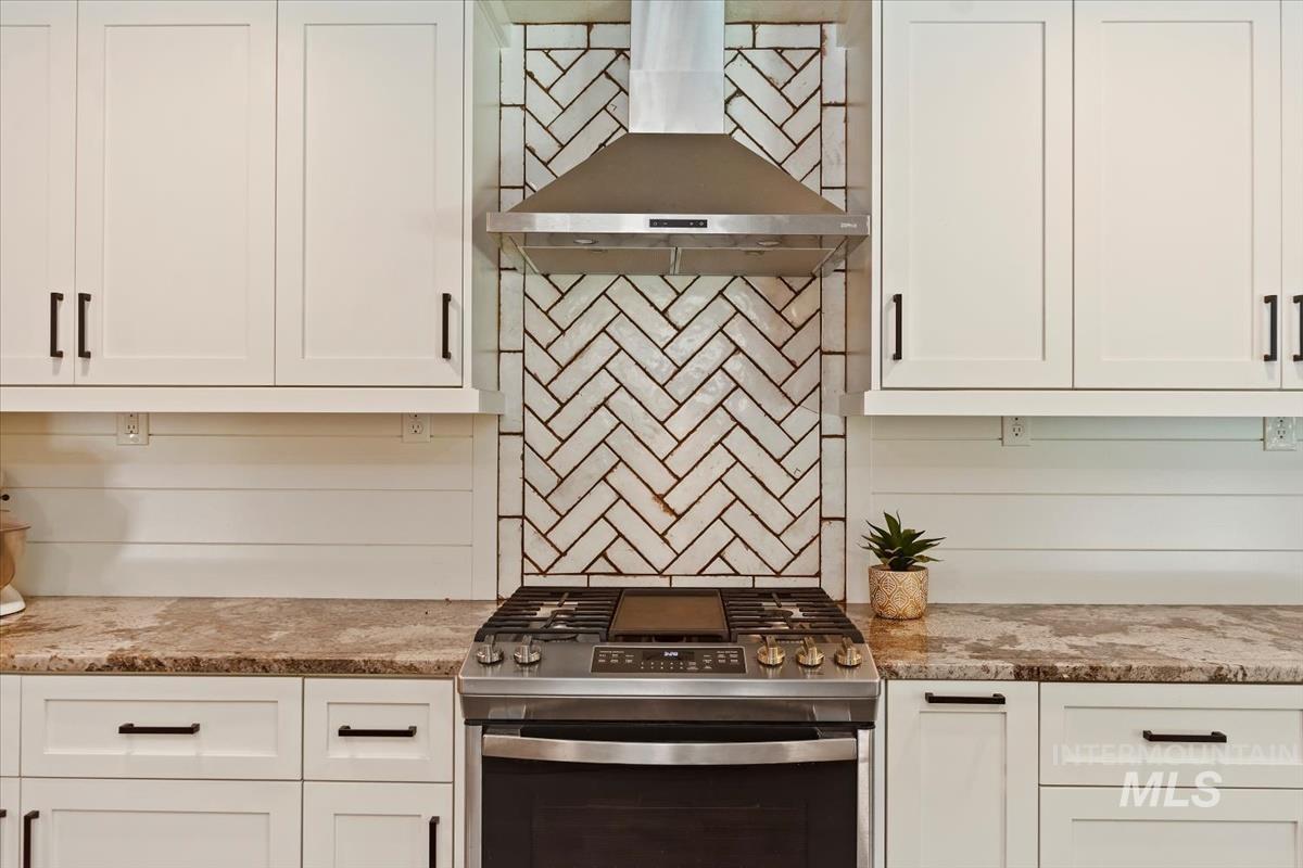 Kitchen featuring stainless steel gas range oven, white cabinets, wall chimney exhaust hood, and light stone countertops