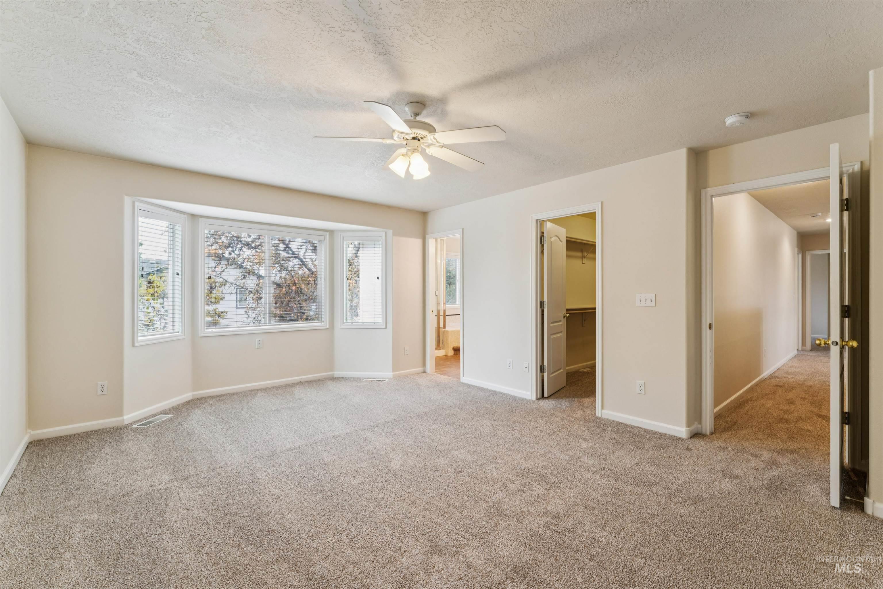 Unfurnished bedroom with light colored carpet, a spacious closet, a textured ceiling, a ceiling fan, and connected bathroom