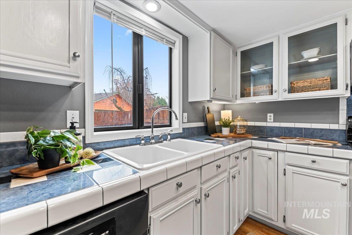 Kitchen with tile counters, white cabinetry, glass insert cabinets, dishwasher, and light wood-style flooring