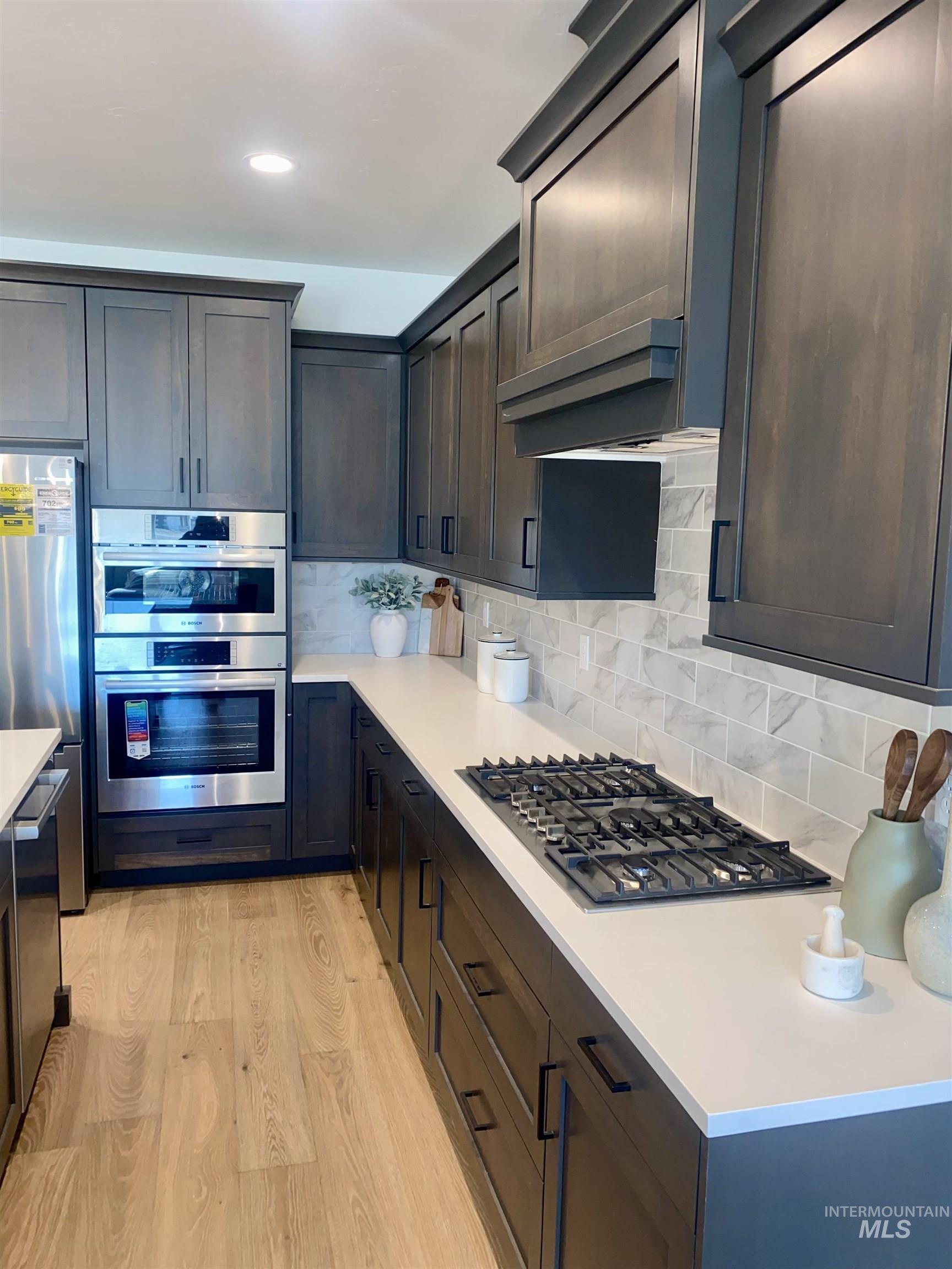Kitchen featuring backsplash, stainless steel appliances, light wood-style flooring, exhaust hood, and light stone counters