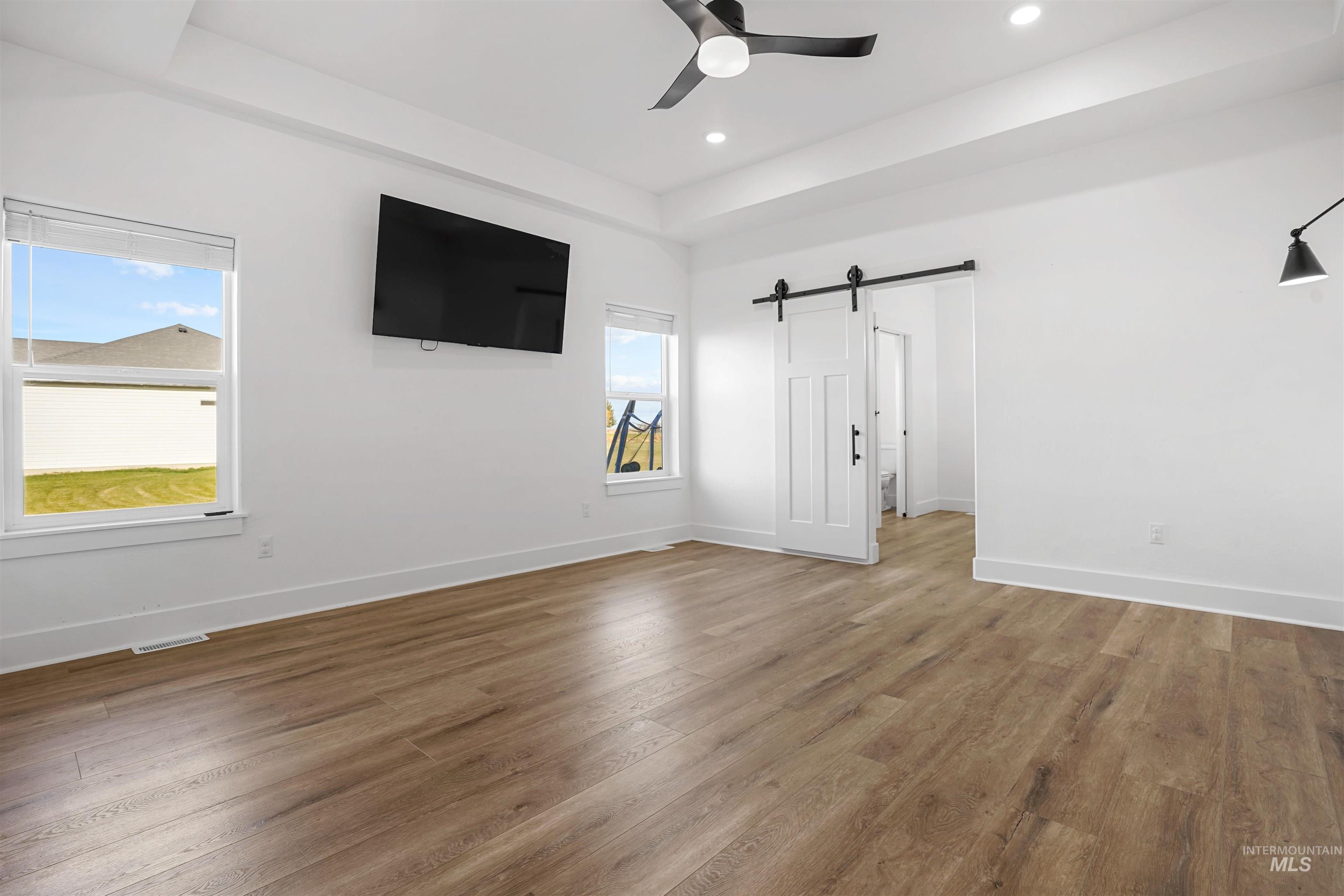 Unfurnished bedroom with a tray ceiling, a barn door, light wood finished floors, a ceiling fan, and recessed lighting