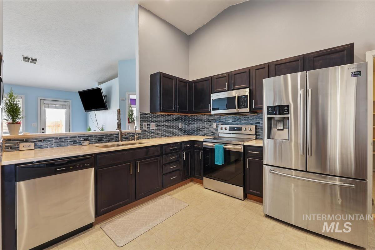 Kitchen featuring stainless steel appliances, decorative backsplash, dark brown cabinets, light flooring, and light stone counters