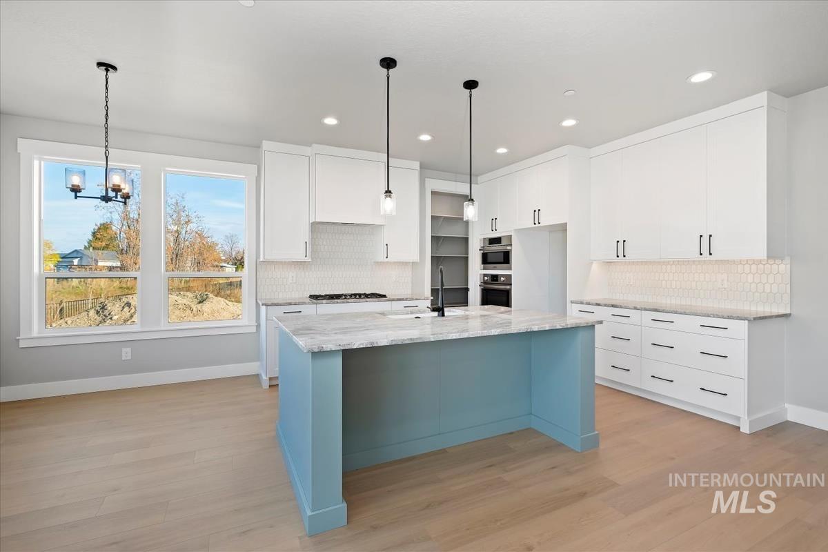 Kitchen featuring decorative light fixtures, white cabinetry, a center island with sink, light stone countertops, and light wood-style floors