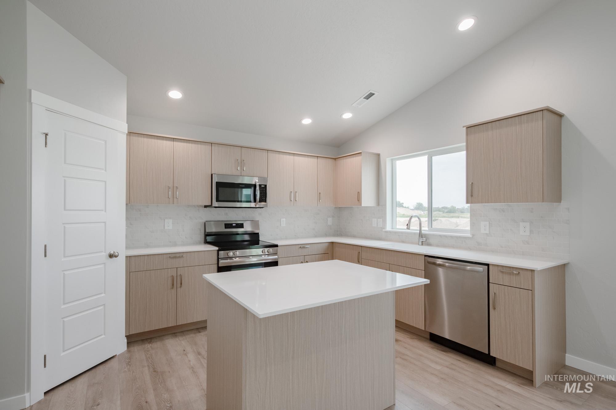 Kitchen with light brown cabinets, decorative backsplash, vaulted ceiling, stainless steel appliances, and light wood-style floors