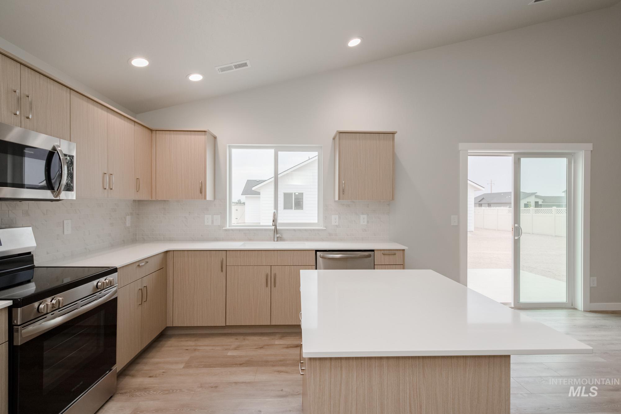 Kitchen with light brown cabinetry, stainless steel appliances, vaulted ceiling, light wood-style flooring, and tasteful backsplash