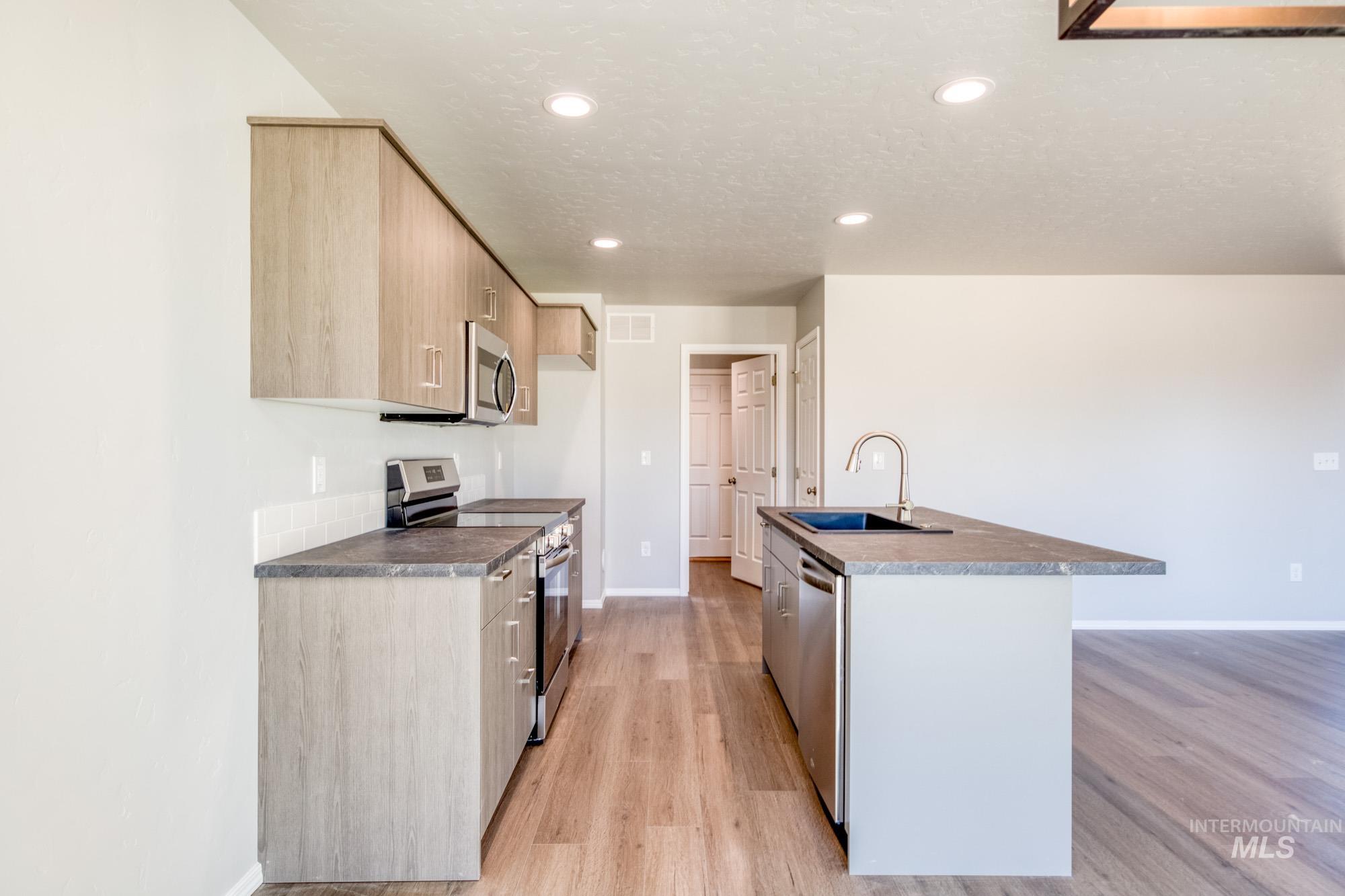 Kitchen featuring appliances with stainless steel finishes, light wood finished floors, an island with sink, recessed lighting, and dark countertops