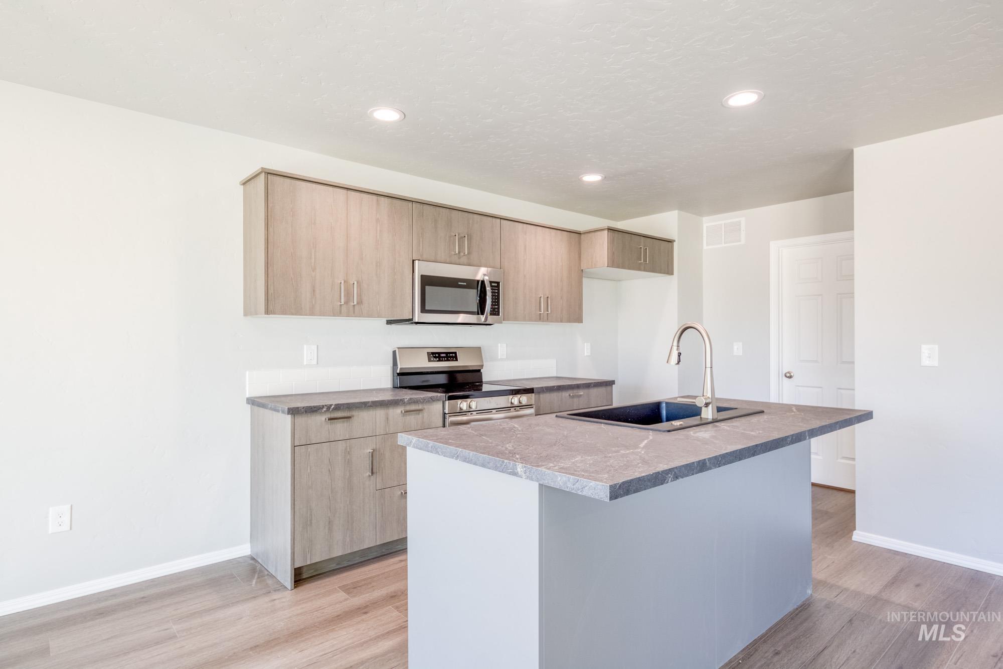 Kitchen with stainless steel appliances, a kitchen island with sink, light brown cabinetry, light wood-type flooring, and recessed lighting